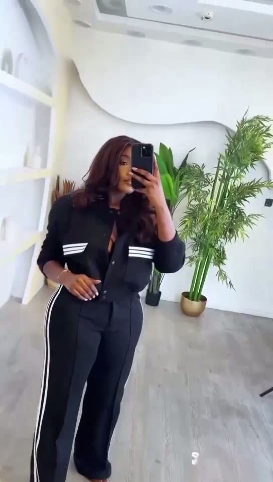 Person wearing a black tracksuit with white stripes taking a mirror selfie in a room with plants.