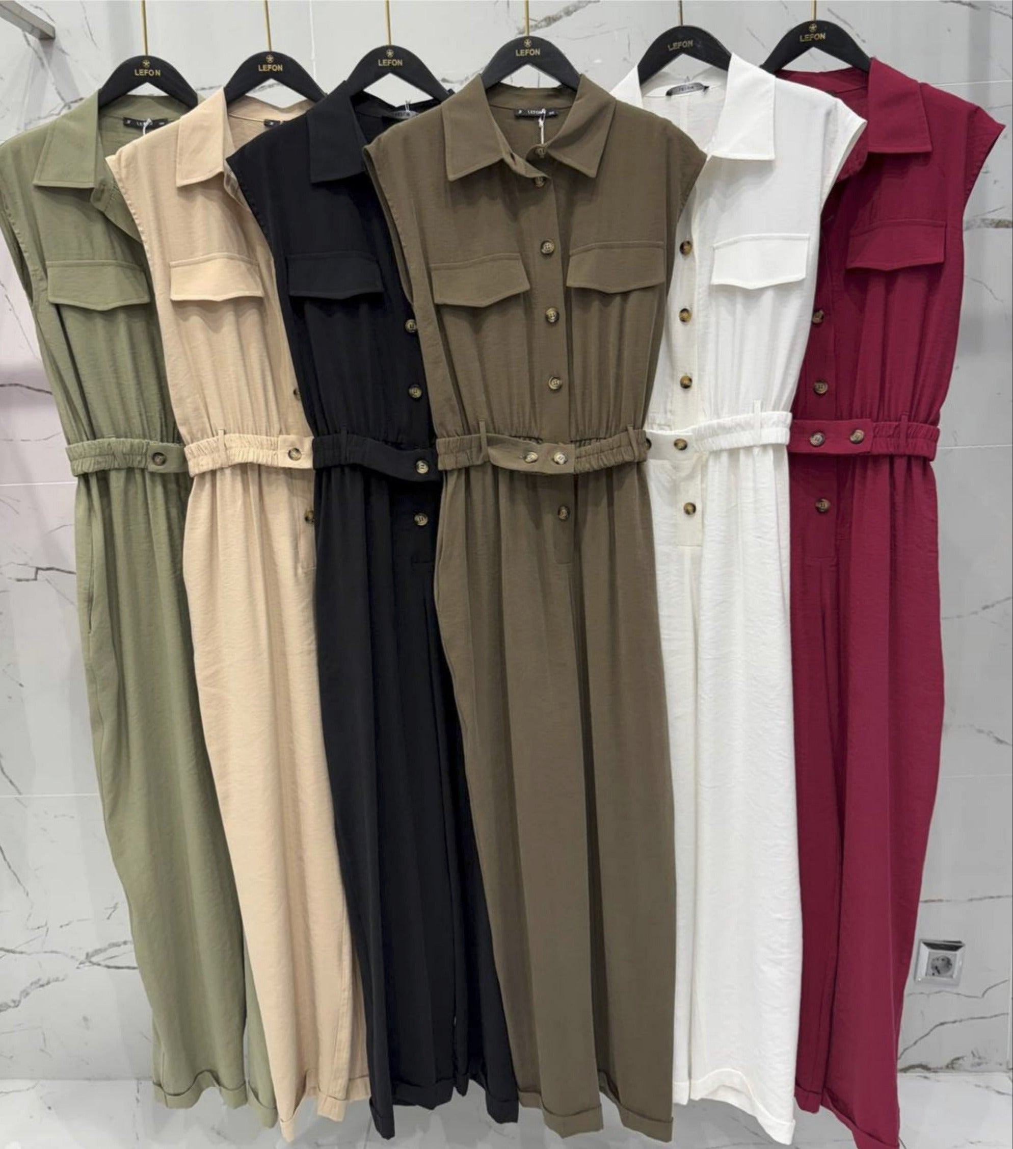 Five different colored jumpsuits hanging on a rack against a marble wall.