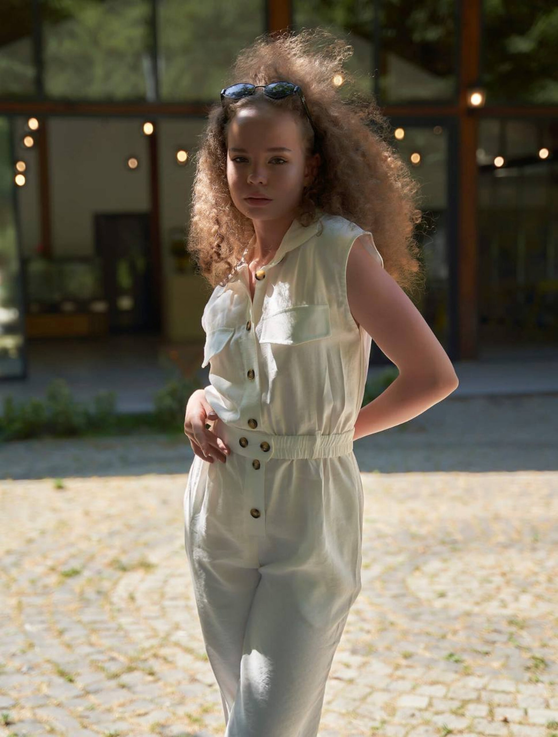 Woman in a light-colored jumpsuit standing outdoors with string lights in the background